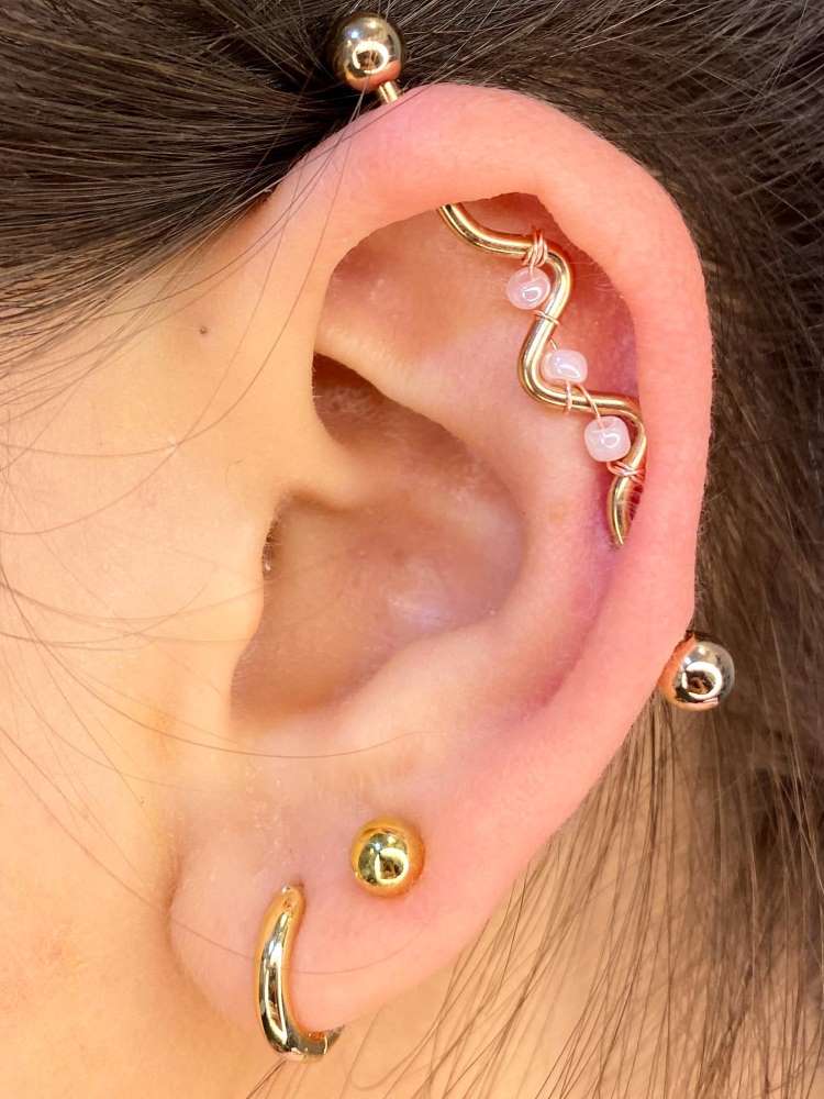 Orelha com piercing em dourado na tranversal, com detalhes em rosa e dois brincos no lóbulo.