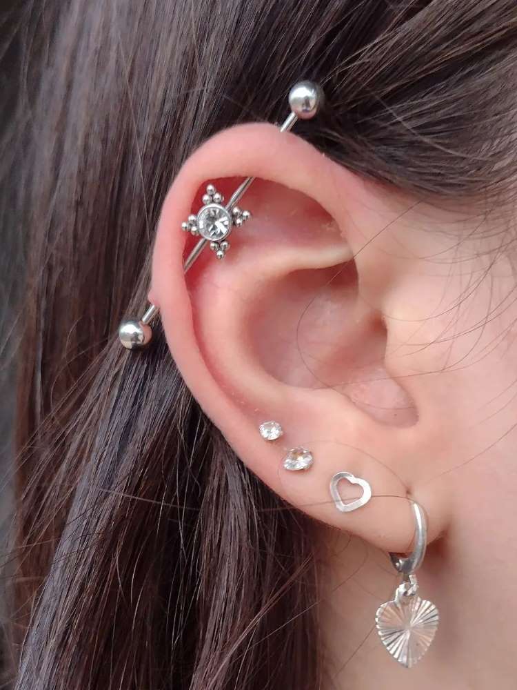 Orelha com piercing transversal com detalhe em estrela e quatro furos no lóbulo, sendo dois pontos de luz, um coração e uma argola com coração, tudo em prata